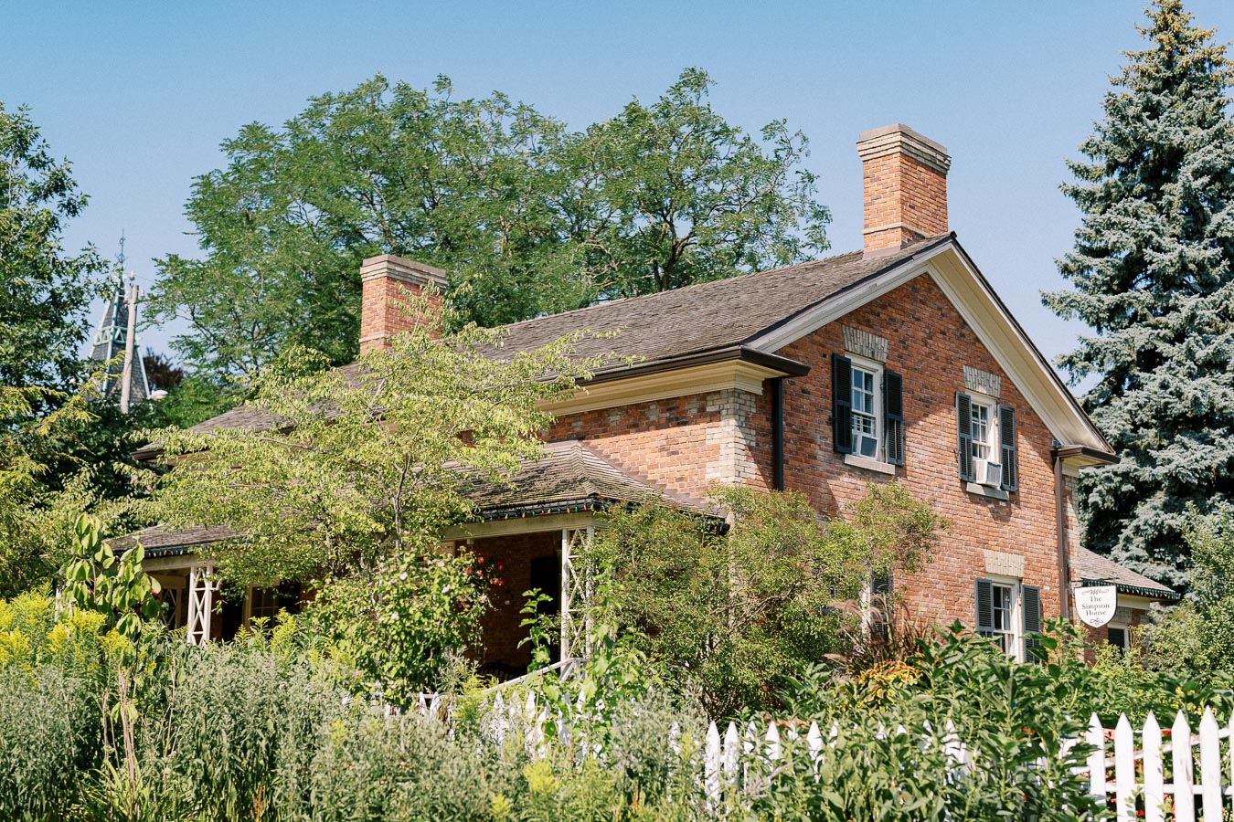 Charming historic brick house surrounded by lush greenery and trees on a sunny day, featuring classic architecture and vintage wooden shutters.