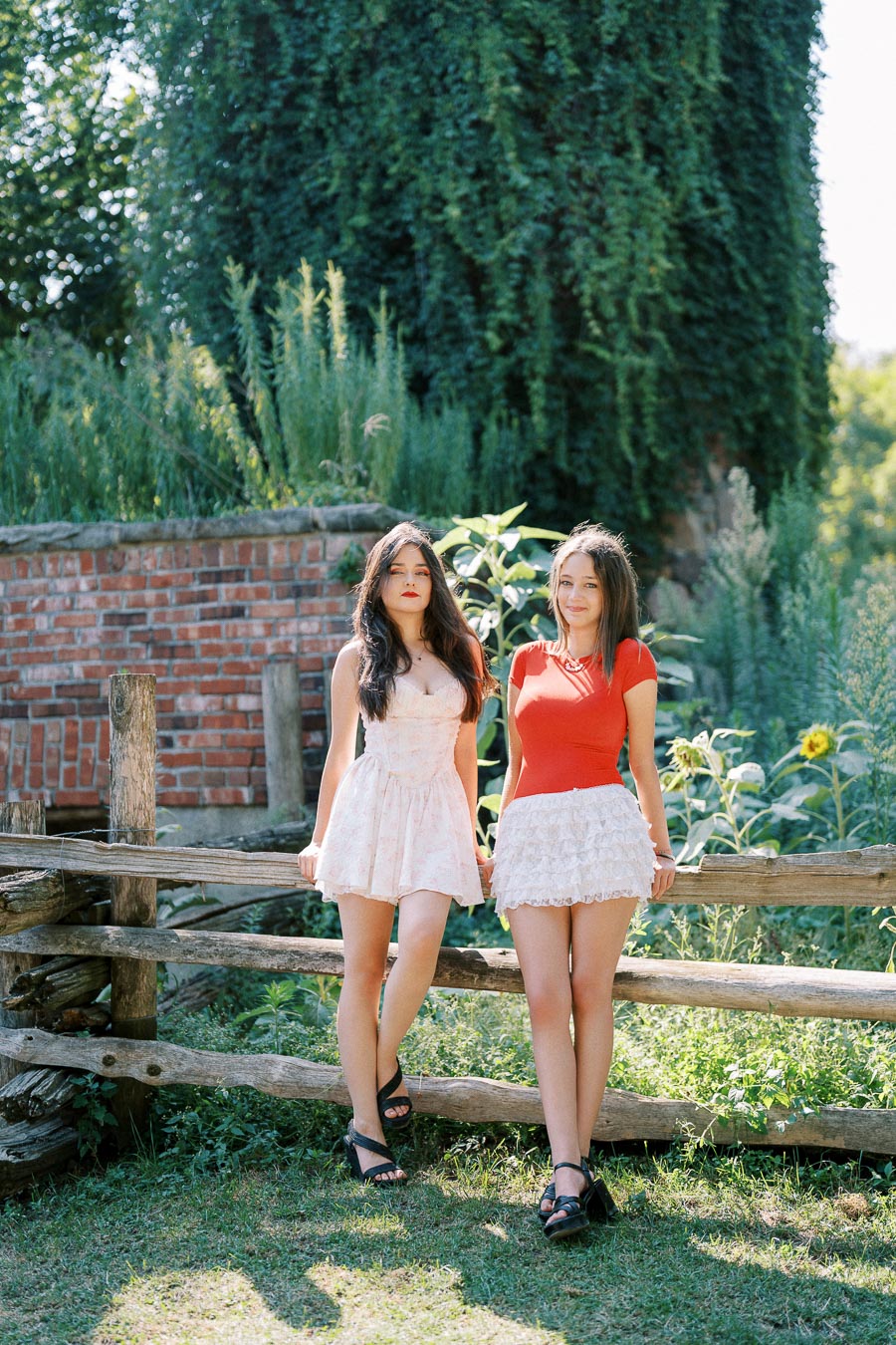 Two women in summer outfits standing in a garden setting near a wooden fence, surrounded by lush green foliage and a brick wall, with sunlight enhancing the scene.
