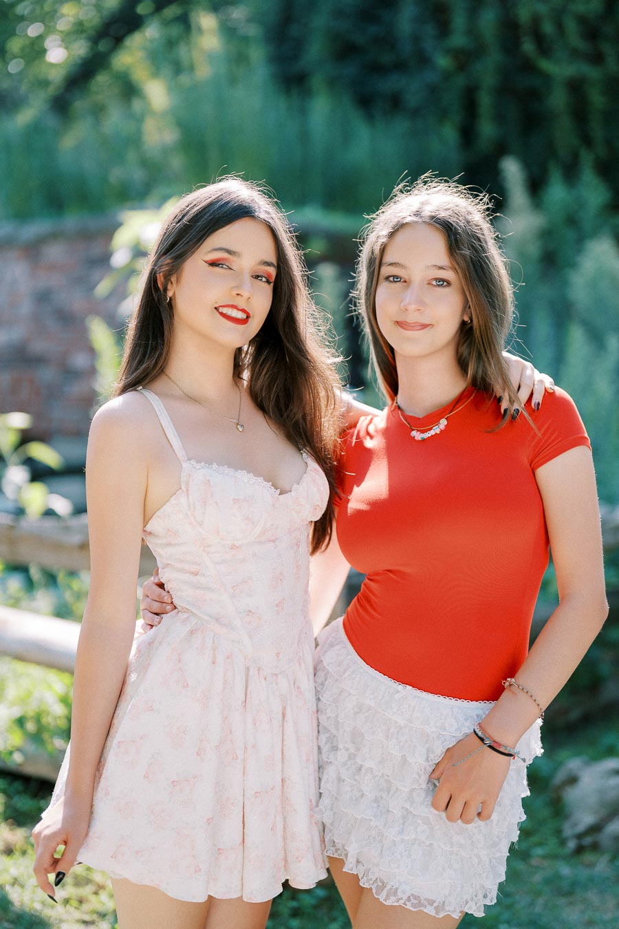 Two young women posing outdoors in summer attire, with one wearing a floral dress and the other in a red top and white lace skirt, standing in a lush green garden setting.
