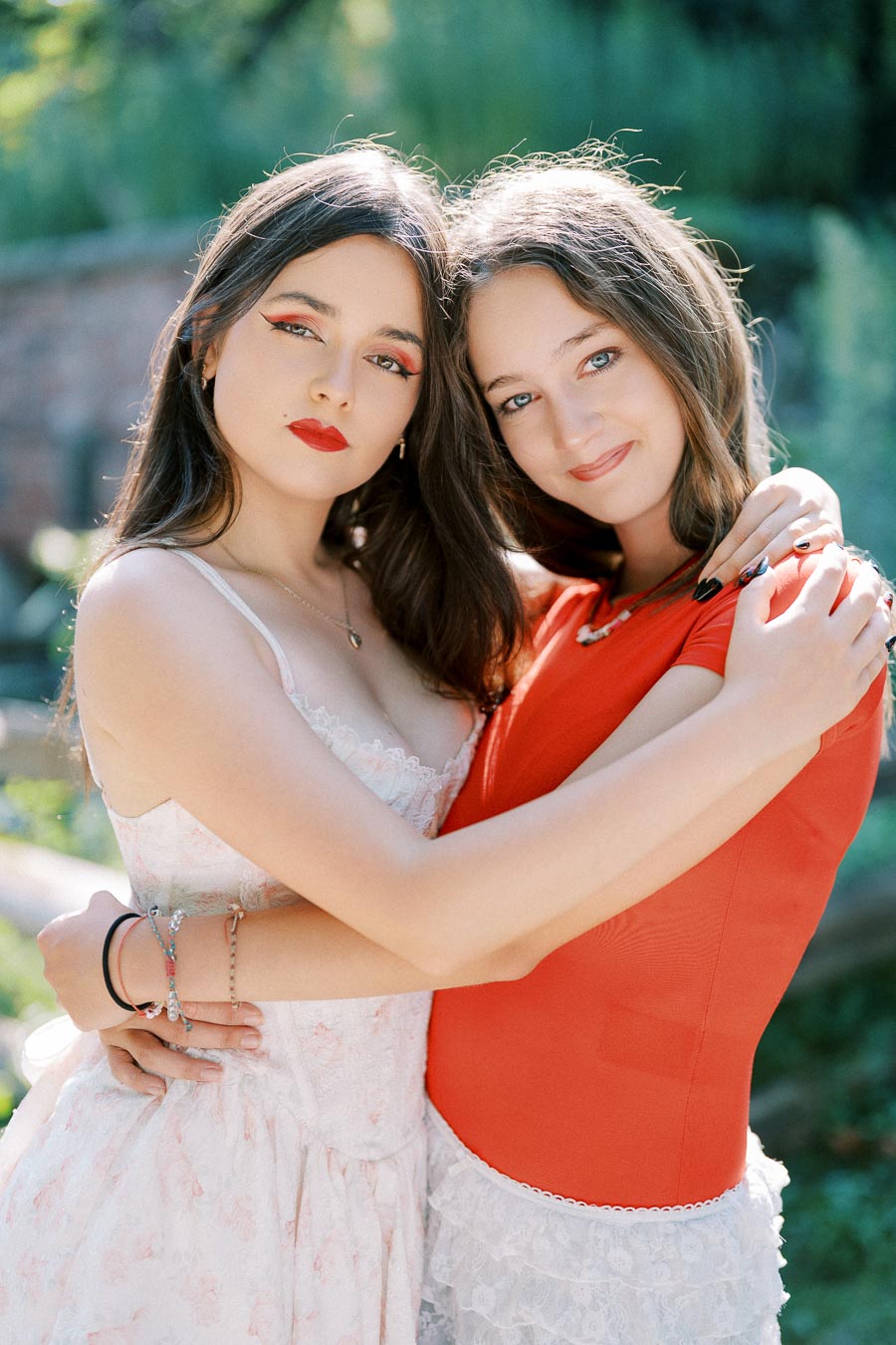 Two young women pose outdoors, embracing each other with smiles, dressed in a white floral dress and a red top, surrounded by lush greenery.