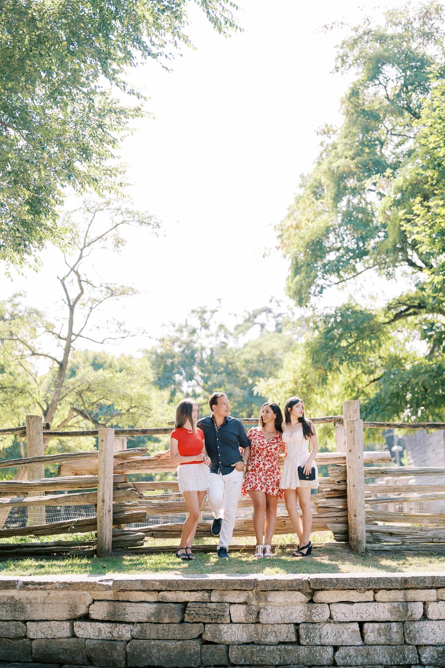 A family enjoying a sunny day outdoors, standing on a stone path in front of a rustic wooden fence. The background features lush green trees under a bright blue sky.