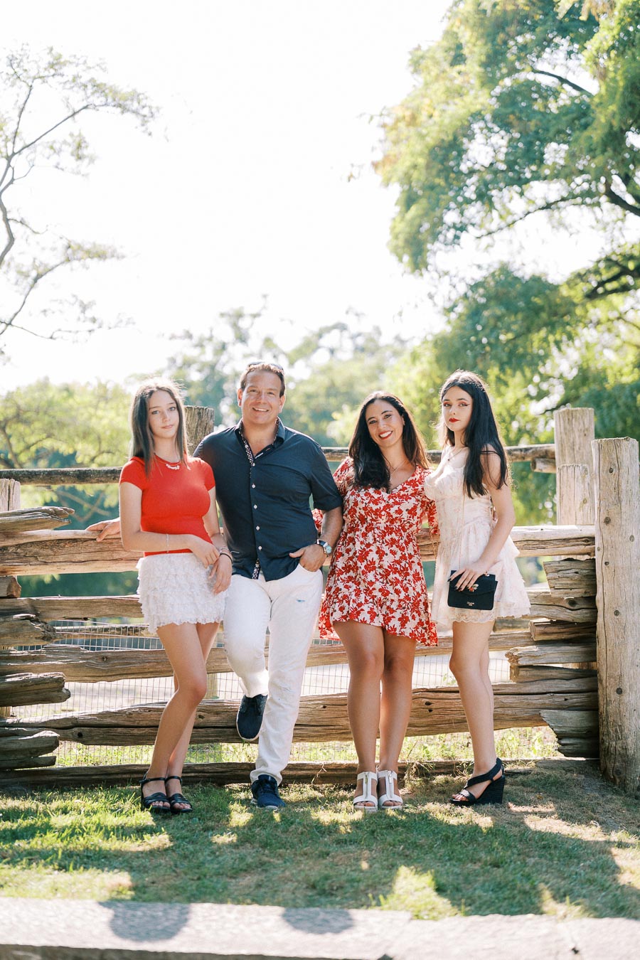 A group of four people standing together outdoors in front of a rustic wooden fence, with trees and sunlight in the background, creating a cheerful and relaxed atmosphere.