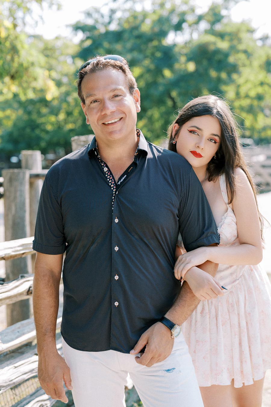 A smiling man in a dark shirt stands outside with a young woman in a light dress leaning on him, surrounded by trees and wooden fence.