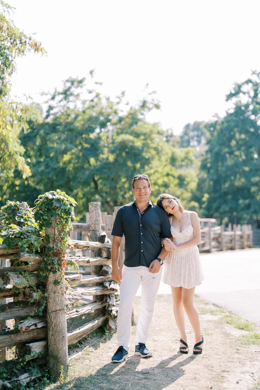 A couple enjoying a sunny day in a park, standing next to a rustic wooden fence with lush greenery in the background. The woman is wearing a light summer dress and leaning affectionately on the man, who is dressed in a dark shirt and white pants.