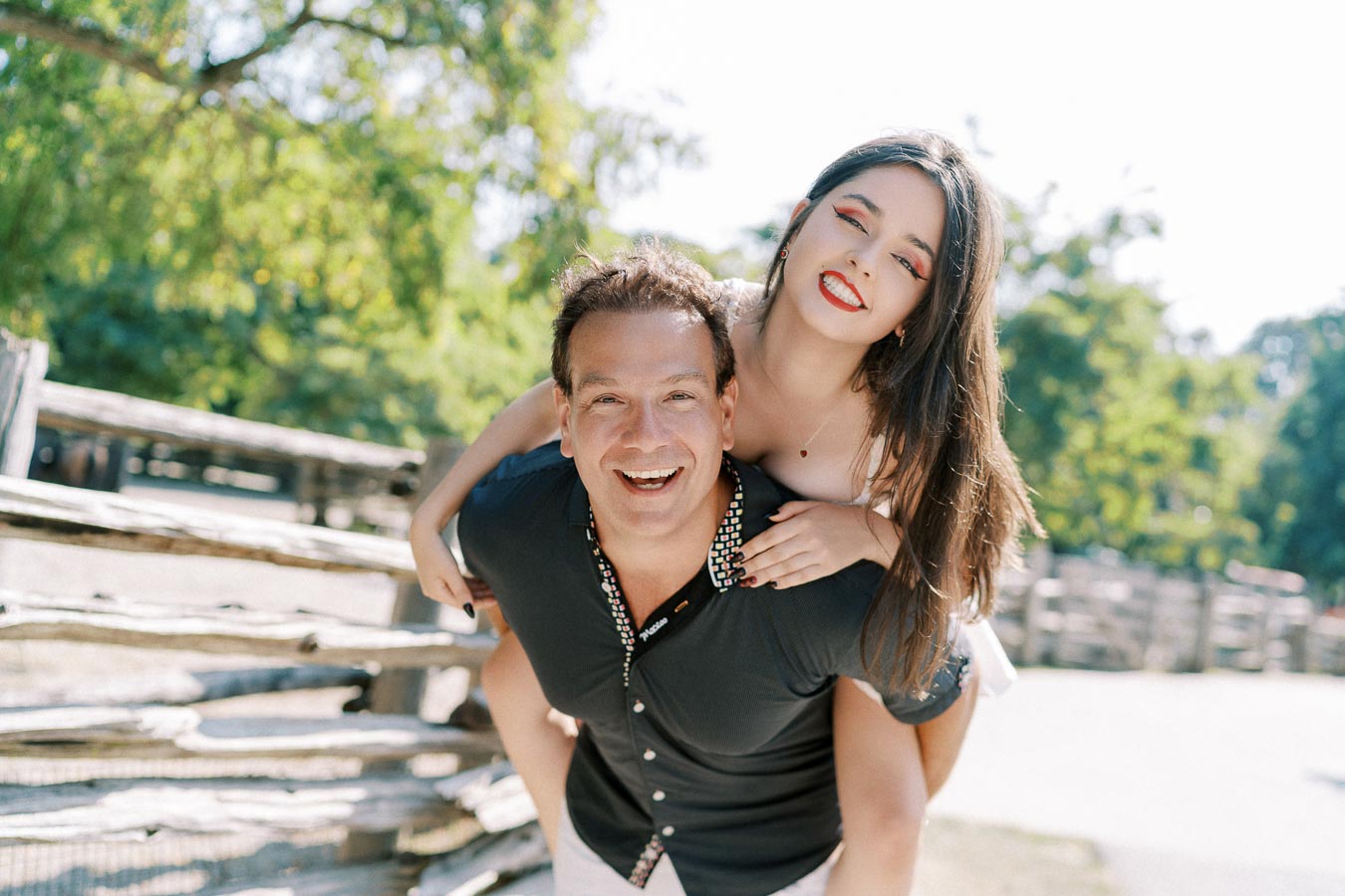 A joyful young man giving a piggyback ride to a smiling woman in a sunny park setting, surrounded by lush green trees and wooden fencing.