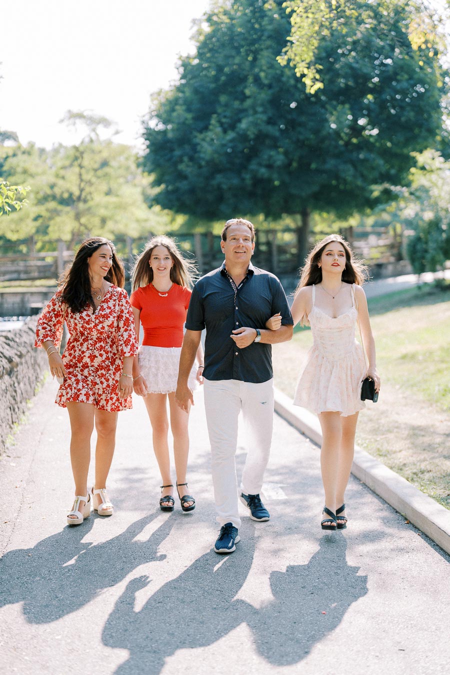 A group of four people walking along a sunny park pathway, with trees and greenery in the background.