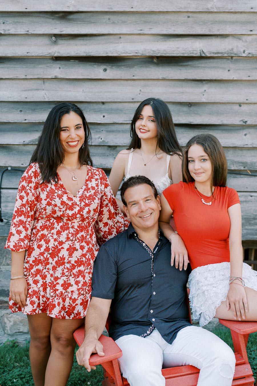 A family of four poses happily outdoors in front of a wooden wall. The mother stands on the left in a red floral dress, the father sits on a red chair wearing a dark shirt and white pants, and their two daughters stand and sit nearby in summer attire, smiling at the camera.