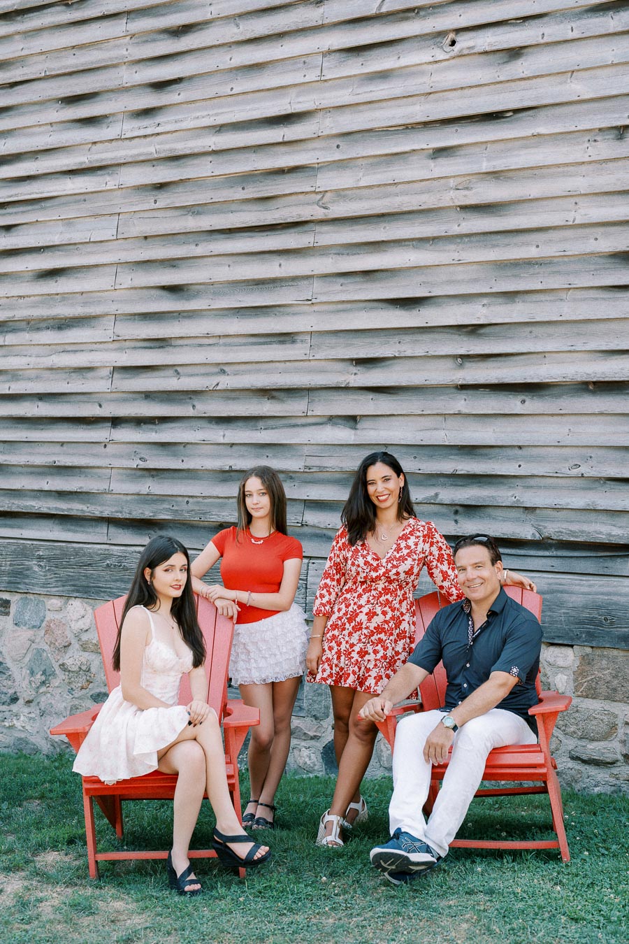 Group of four people in colorful outfits posing outdoors on red chairs in front of a rustic wooden wall.