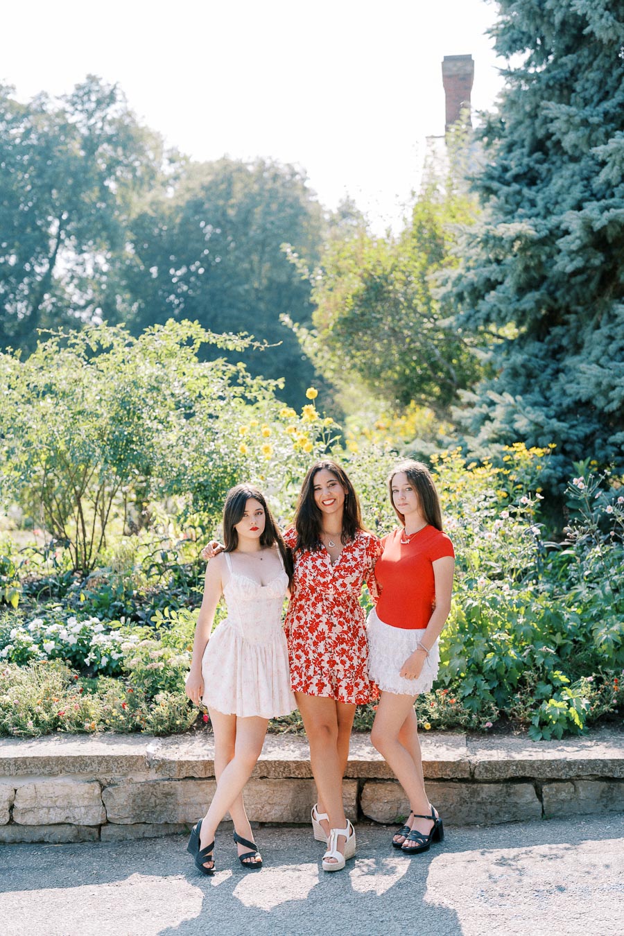 Three women in stylish summer dresses posing outdoors in a sunny garden with lush greenery and flowers.