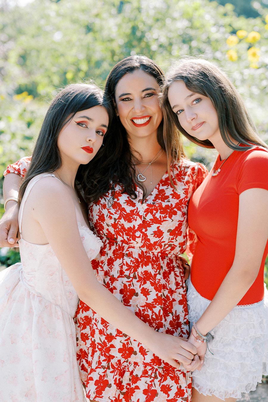 Three women standing closely together outdoors, wearing red and white floral dresses, smiling and holding hands amidst a green, sunny background.