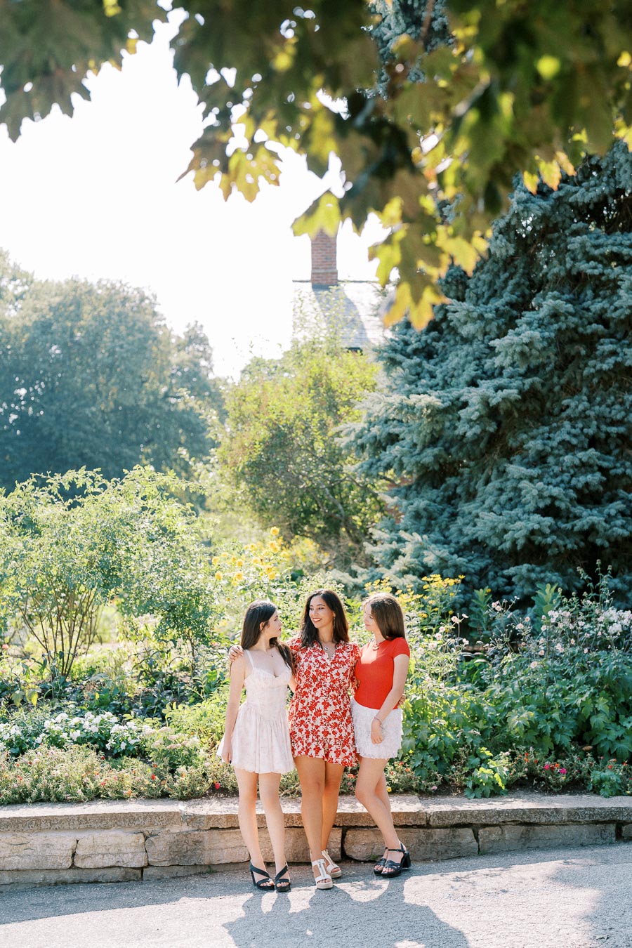 Three women in summer dresses smiling and enjoying a sunny day in a lush garden, surrounded by vibrant greenery and flowers.