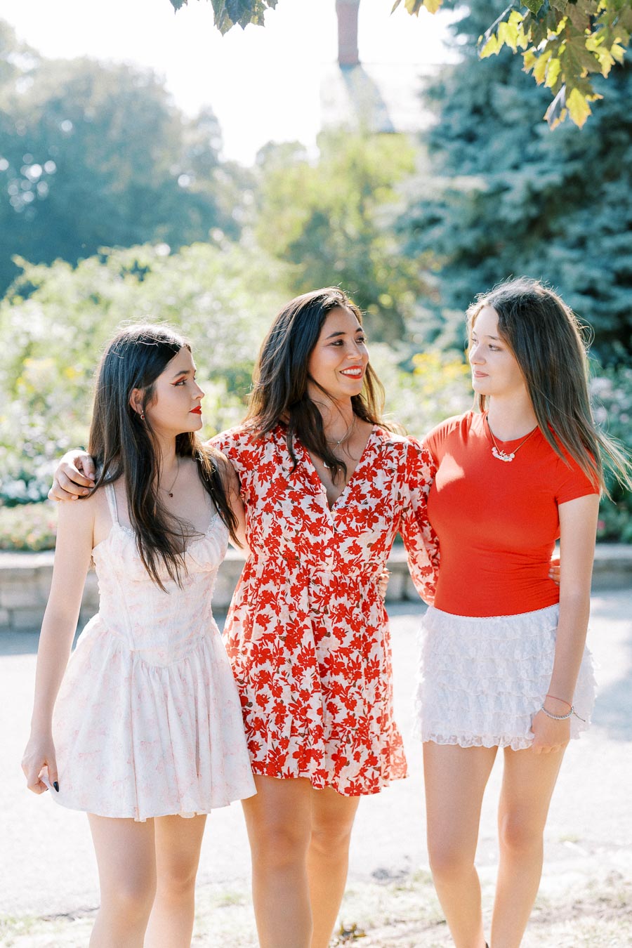 Three young women walking together in a park on a sunny day, wearing stylish summer dresses and smiling at each other, surrounded by lush greenery.