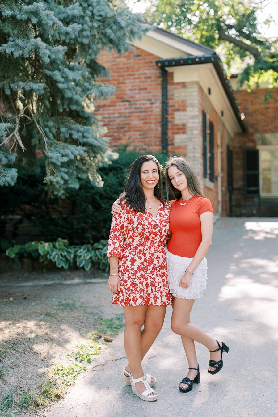 Two women smiling and posing together outdoors by a brick building, with one wearing a red floral dress and the other in a red top and white skirt, surrounded by greenery and sunlight.