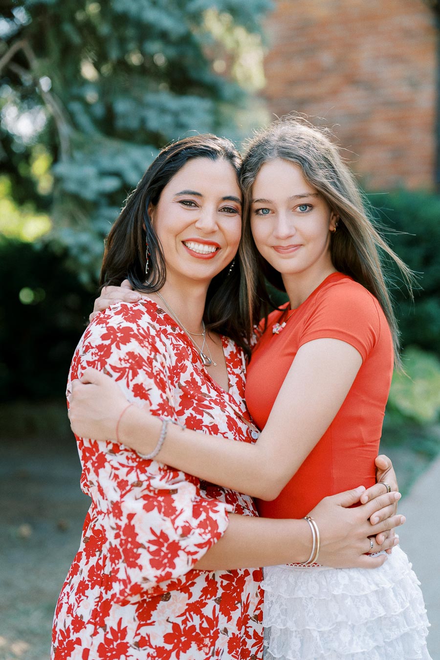 Two women smiling and embracing outdoors, wearing red and white attire, with green foliage in the background.