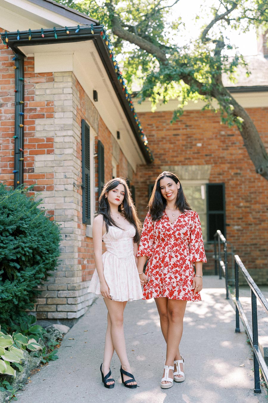 Two women standing outdoors in summer dresses, one in a white and the other in a red floral pattern, posing in front of a brick building with lush greenery and a tree in the background.