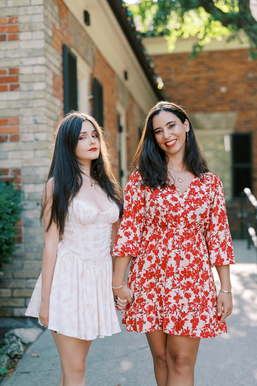 Two women holding hands and smiling outdoors, dressed in stylish floral dresses, standing in a sunlit area with a brick building in the background.
