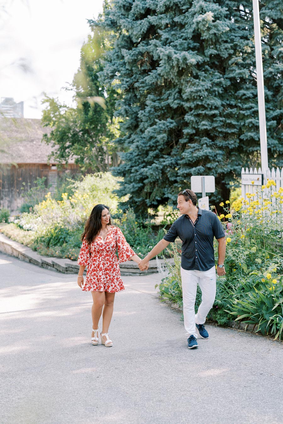 A couple holding hands and walking along a garden path with lush green trees and blooming flowers on a sunny day. The woman is wearing a red floral dress, while the man is dressed in a dark shirt and white pants, evoking a sense of romance and summer bliss.