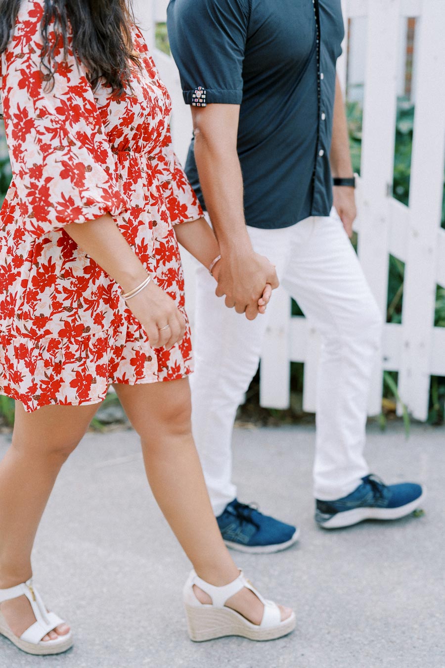 A couple holding hands while walking, with the woman wearing a red floral dress and the man in a dark blue shirt and white pants, walking past a white picket fence.