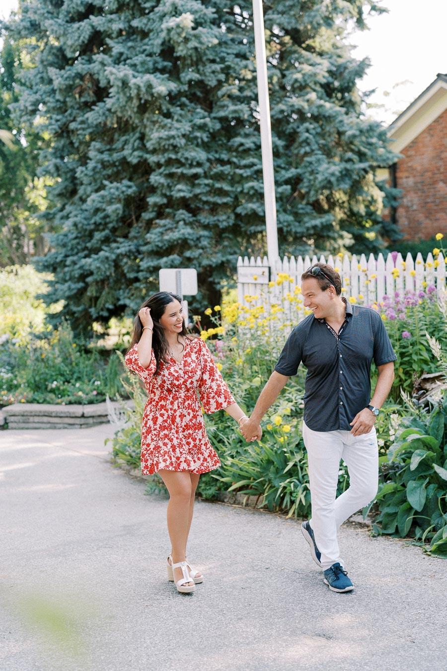 A happy couple holding hands and walking in a garden path, surrounded by lush green plants and vibrant yellow flowers, under a clear blue sky.