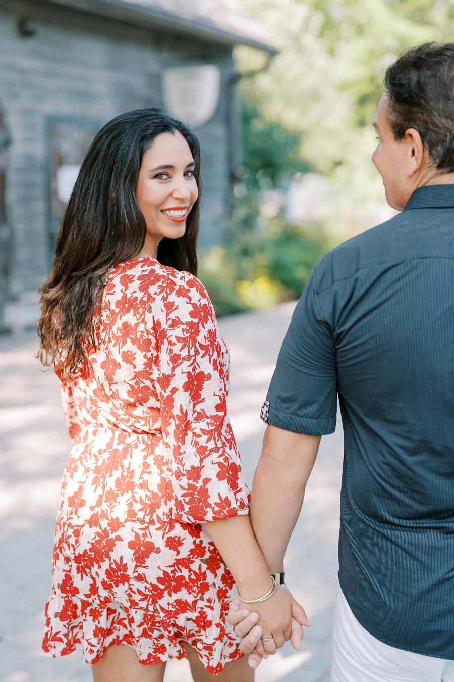 A smiling woman in a red floral dress holding hands with a man in a blue shirt, walking outdoors on a sunny day.