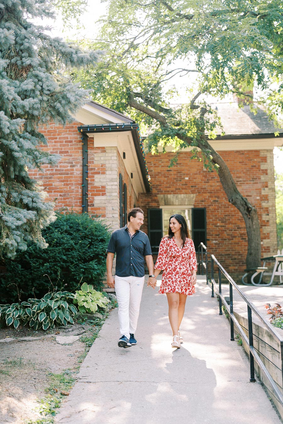 A couple holding hands and walking down a garden path in front of a charming brick building, surrounded by lush greenery.
