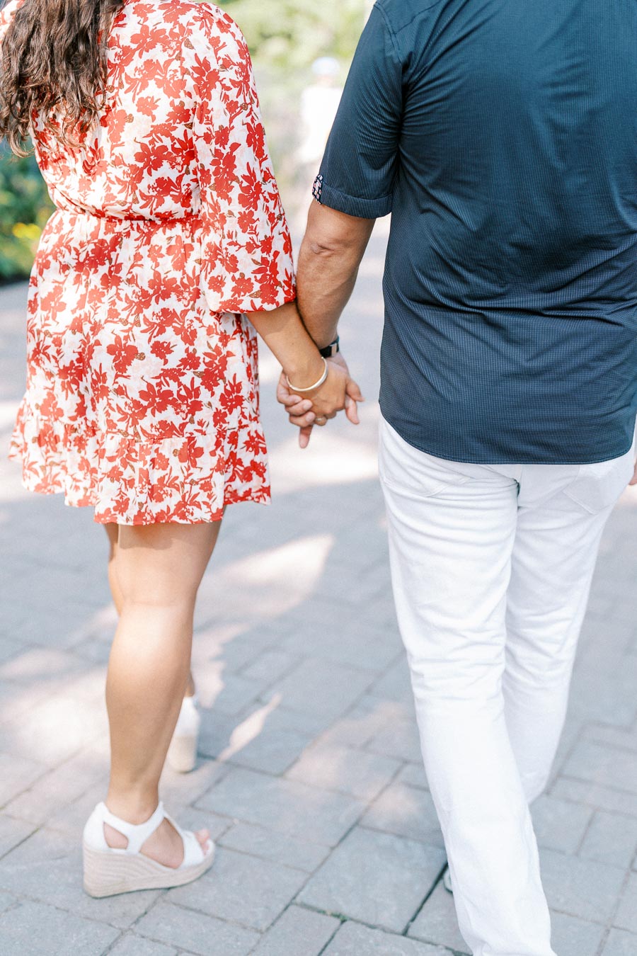 A couple walking hand in hand on a sunlit pathway, with the woman in a red floral dress and the man in a navy shirt and white pants, capturing a romantic moment.