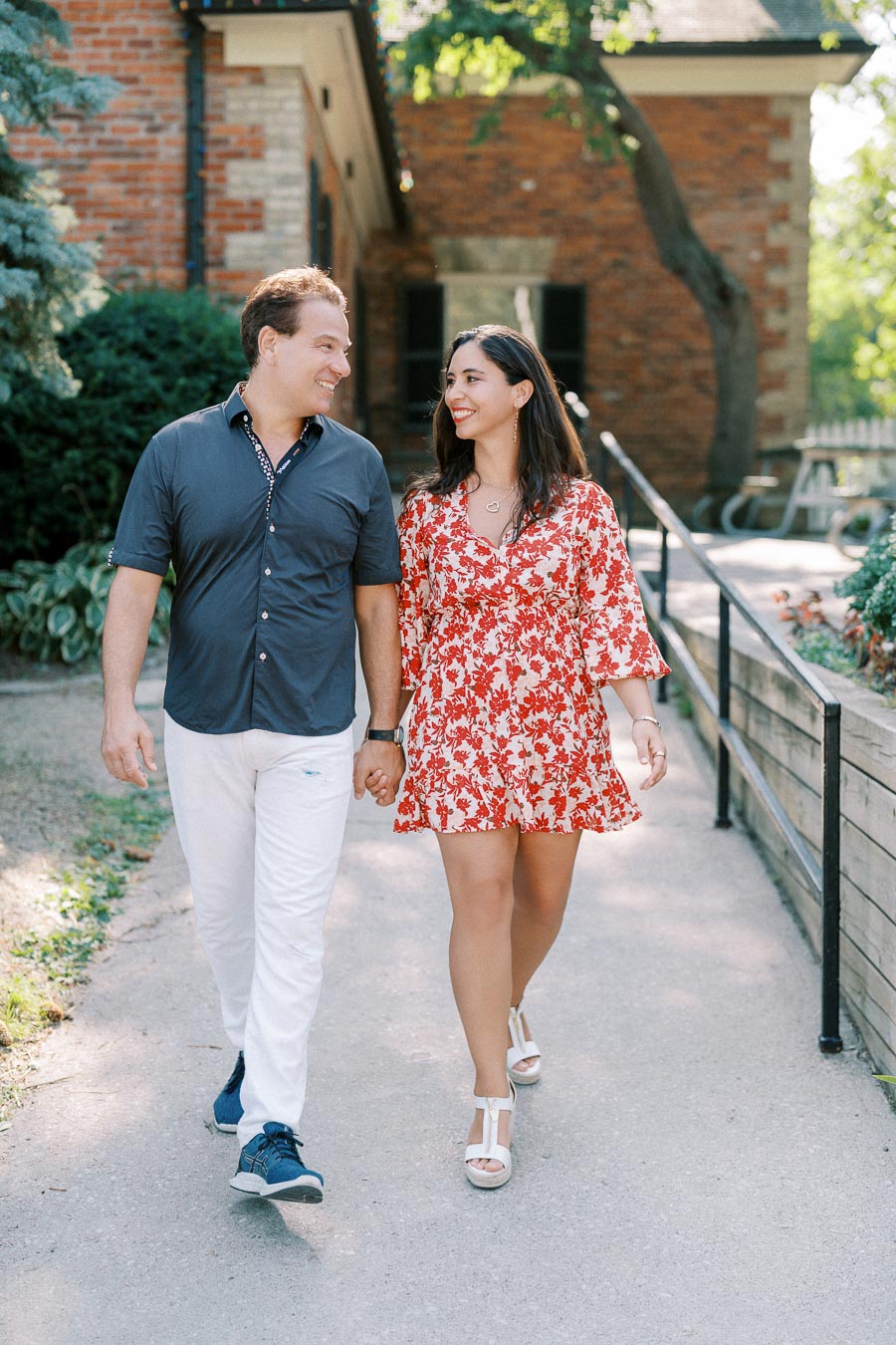 Couple walking hand in hand on a sunlit path in front of a brick house, both smiling and enjoying a casual outdoor stroll.