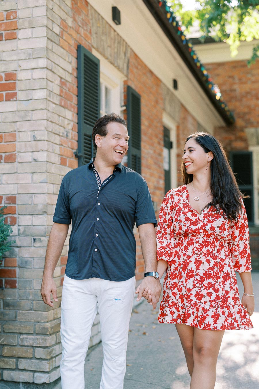 Happy couple holding hands and walking in front of a brick building, smiling at each other on a sunny day.