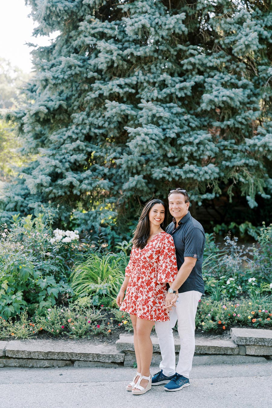 Couple smiling in a park with lush greenery and a large tree in the background, enjoying a sunny day.