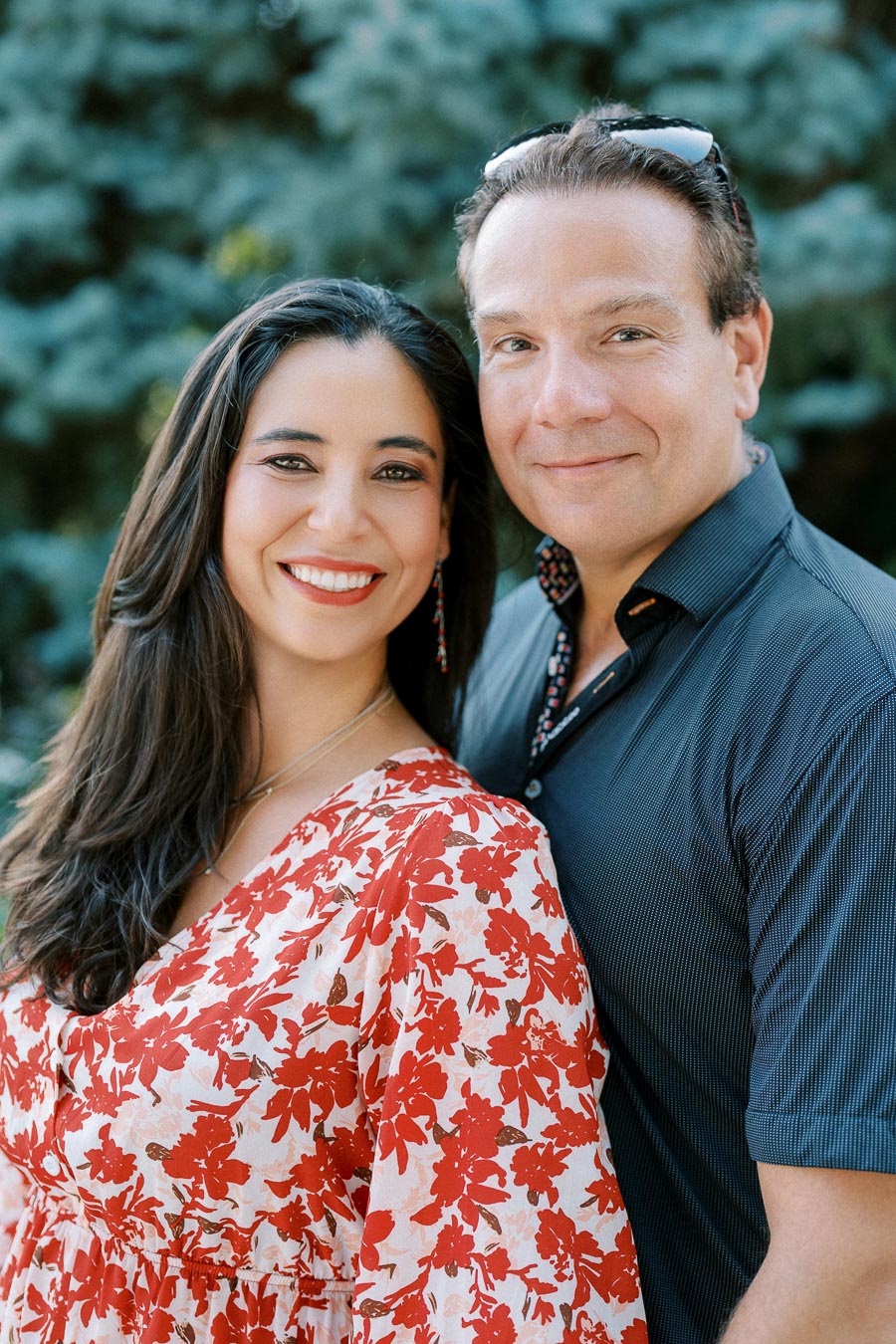 Smiling couple posing outdoors in floral attire, surrounded by lush greenery.