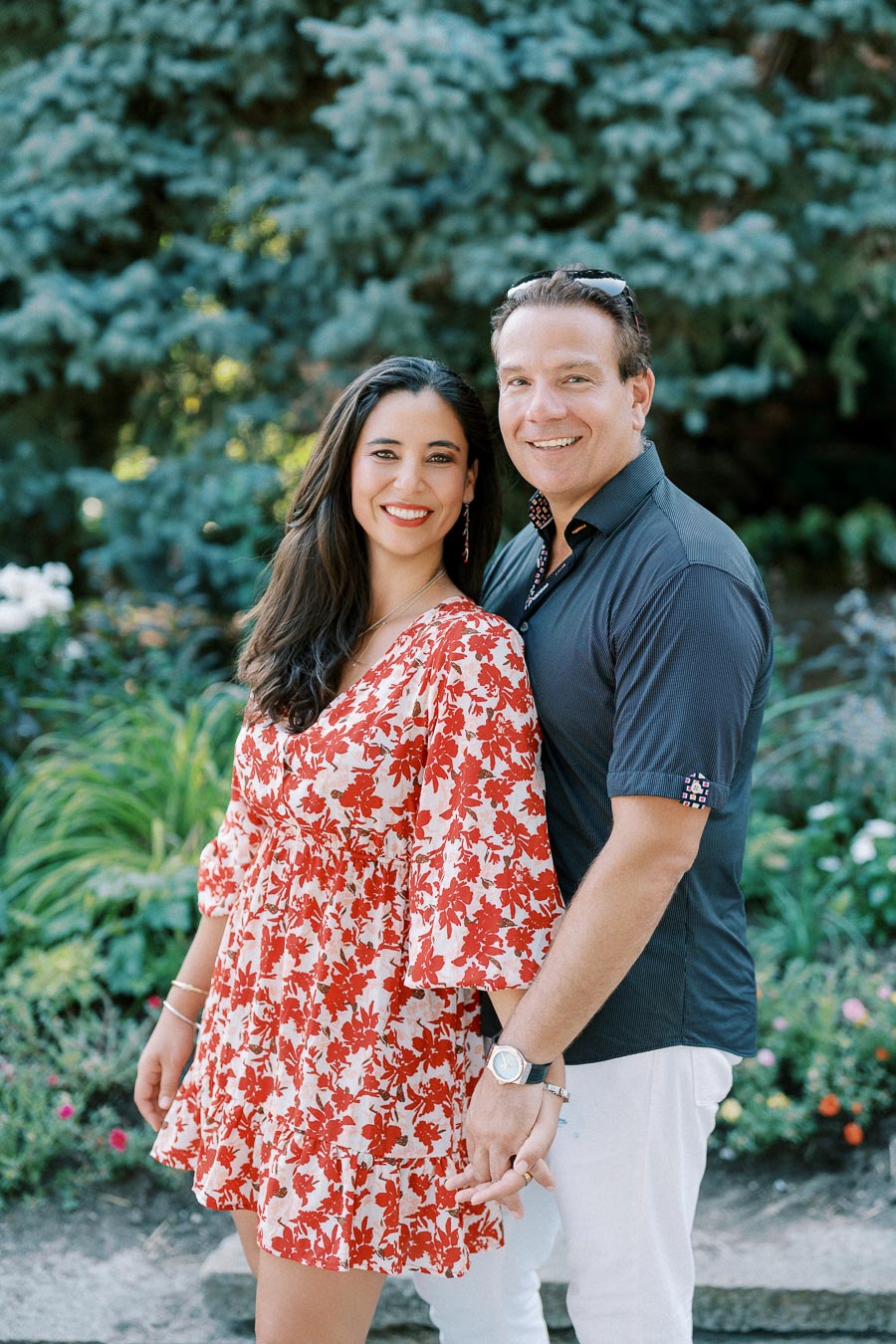 A happy couple enjoying a sunny day in a lush garden setting, with the woman wearing a red and white floral dress and the man in a navy shirt and white pants, both smiling and holding hands.