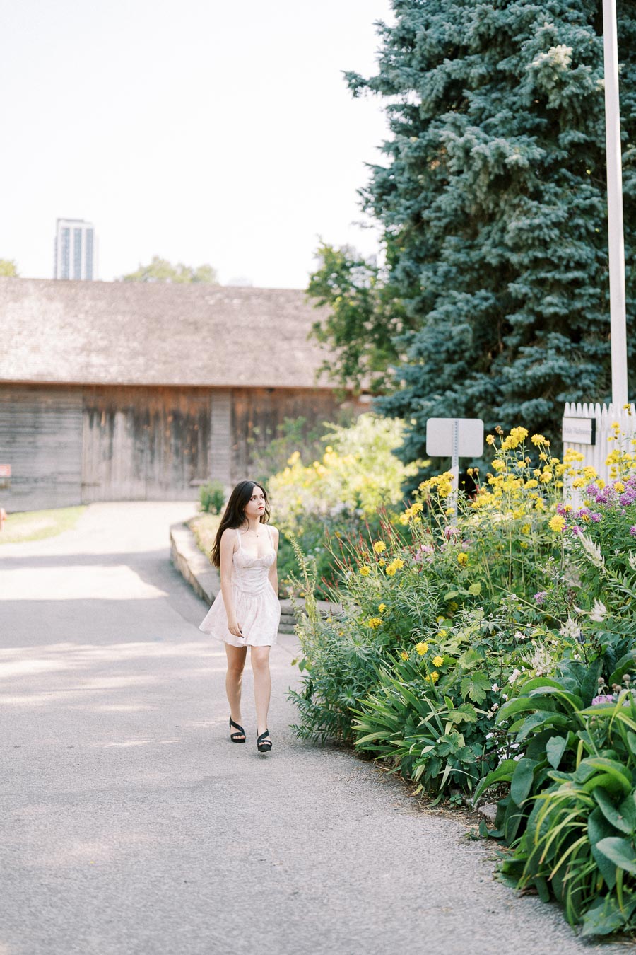 Young woman in a white dress walking along a garden path lined with vibrant yellow and purple flowers, with a rustic wooden building and tall green trees in the background.