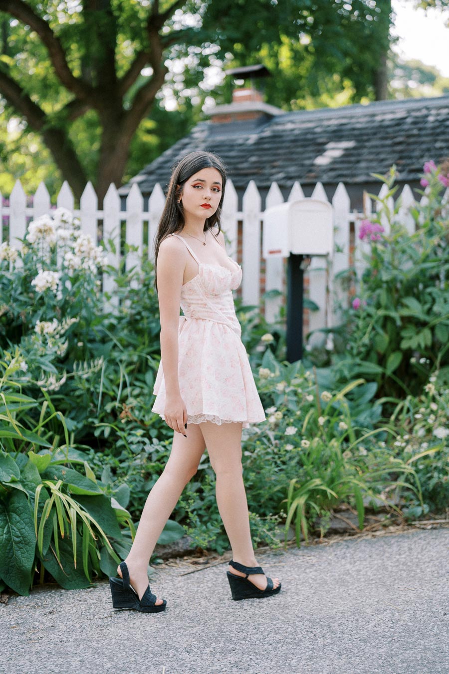 A young woman in a light pink summer dress walks along a garden path with lush greenery and a white picket fence in the background, embodying a serene, summery vibe.