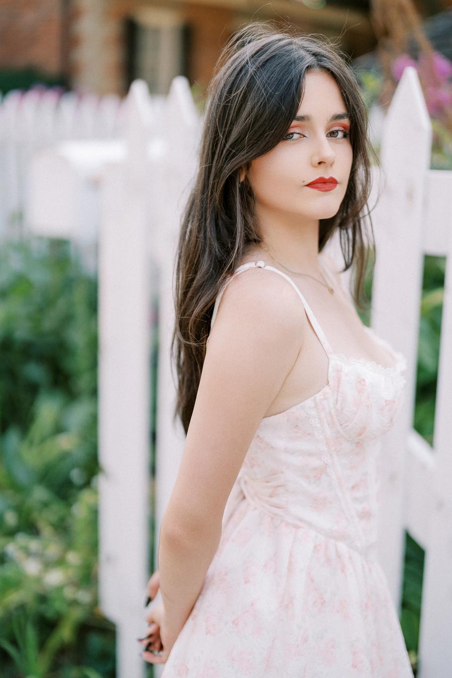 A young woman in a floral dress stands beside a white picket fence, surrounded by lush greenery and flowers, exuding a serene and elegant vibe.