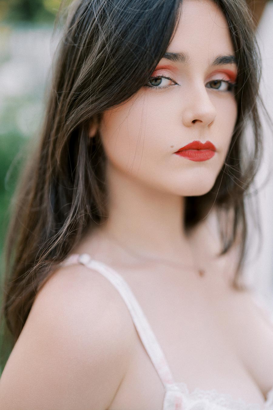 Portrait of woman with long dark hair and red lipstick, wearing a white top in an outdoor setting.