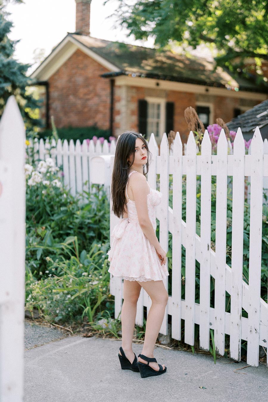 A young woman in a light floral dress posing near a white picket fence with a brick house and garden in the background.