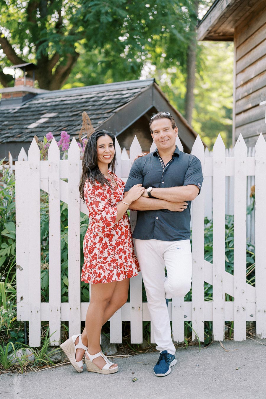 ```plaintext Happy couple standing in front of a white picket fence, posing outdoors on a sunny day. The woman is wearing a red floral dress and sandals while the man wears a dark shirt and white pants. ```