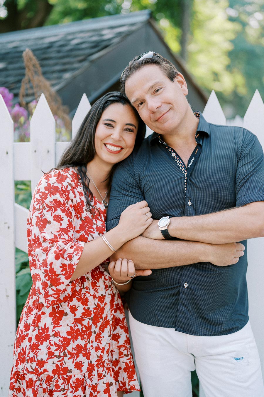 A happy couple standing in front of a white picket fence, the woman in a red floral dress and the man in a navy shirt, embracing and smiling in a garden setting.