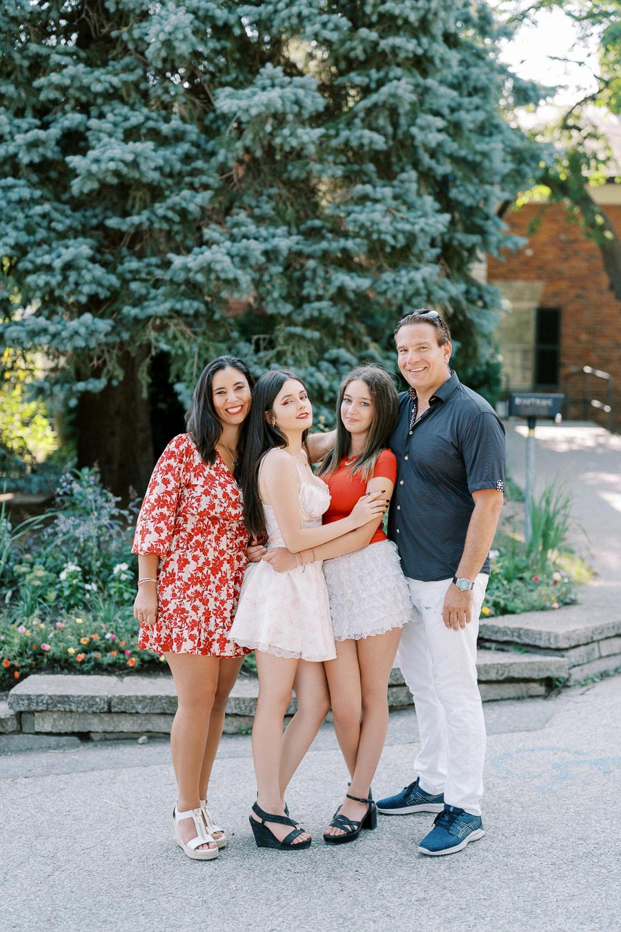 A family of four smiling and posing together in a garden, with a large tree and flowers in the background, creating a joyful and natural outdoor setting.