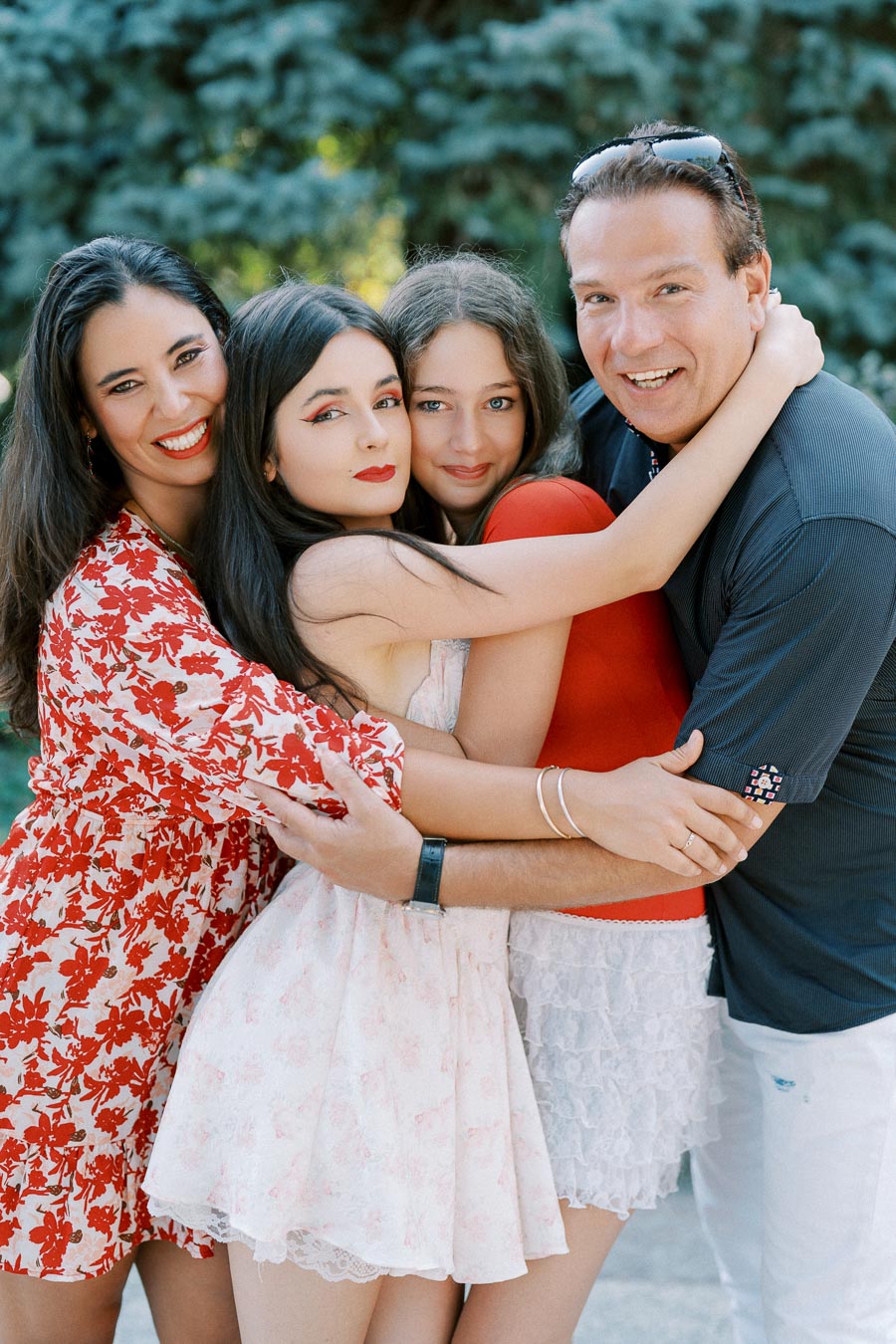 A happy family of four, including a mother in a floral dress, two daughters in light dresses, and a father in a dark shirt, embracing outdoors on a sunny day with greenery in the background.