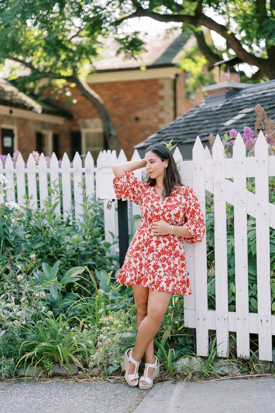 Young woman in a red floral dress poses casually against a white picket fence in front of a charming garden and brick house under lush green trees.
