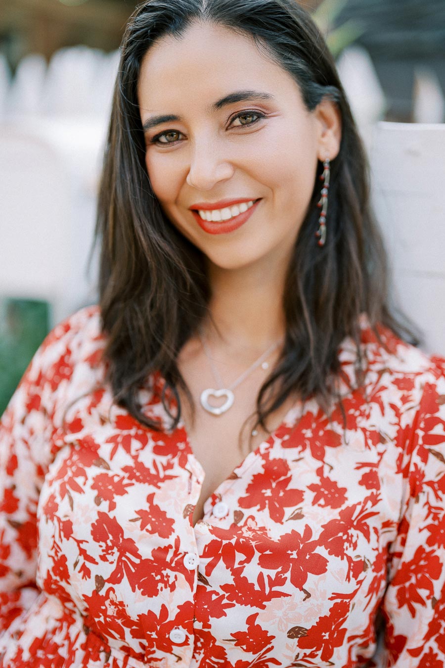 Smiling woman with long dark hair wearing a floral red and white dress, accessorized with heart-shaped necklace and earrings.