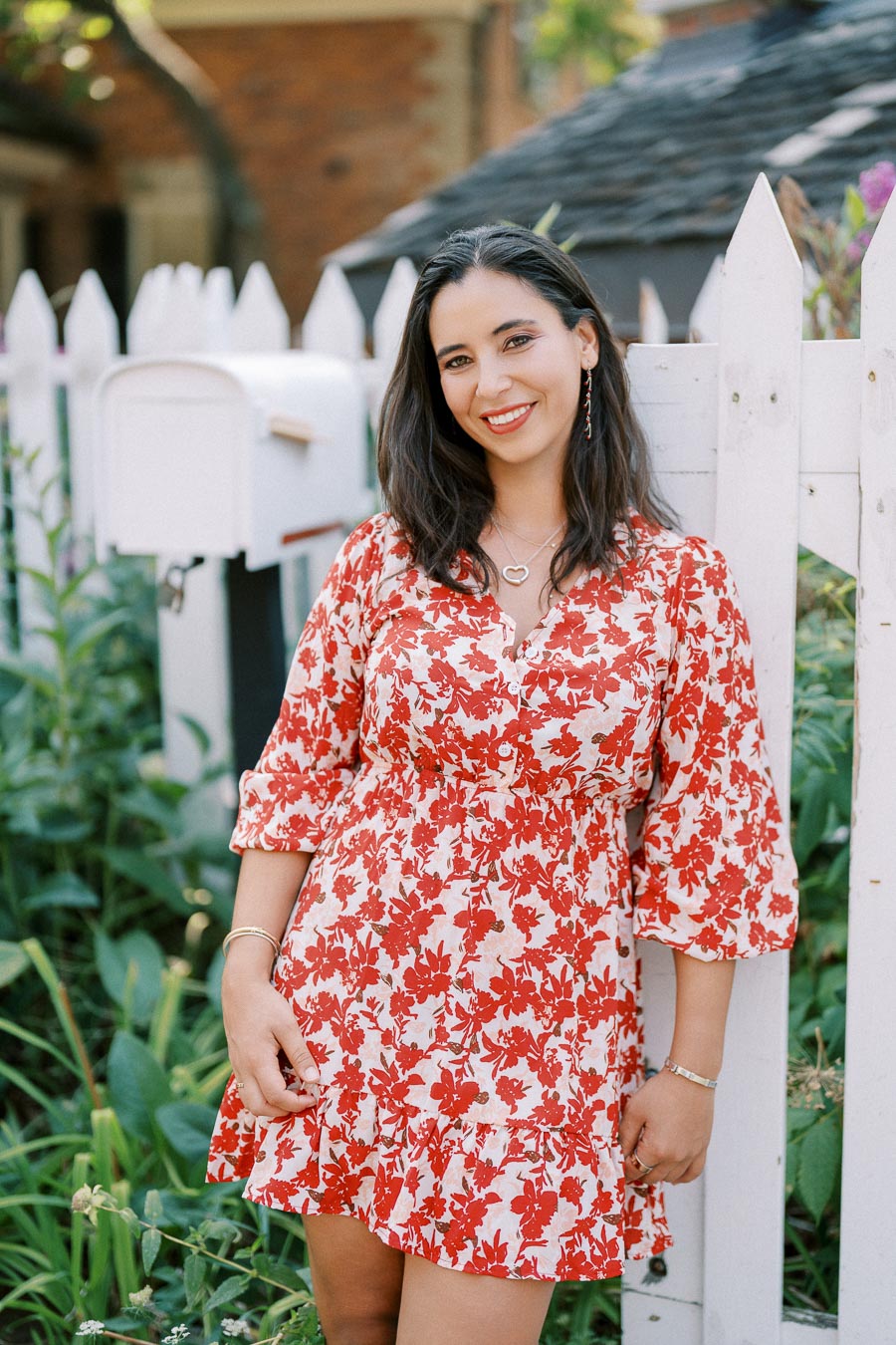 A woman in a red and white floral dress smiles while standing in front of a white picket fence near a garden.