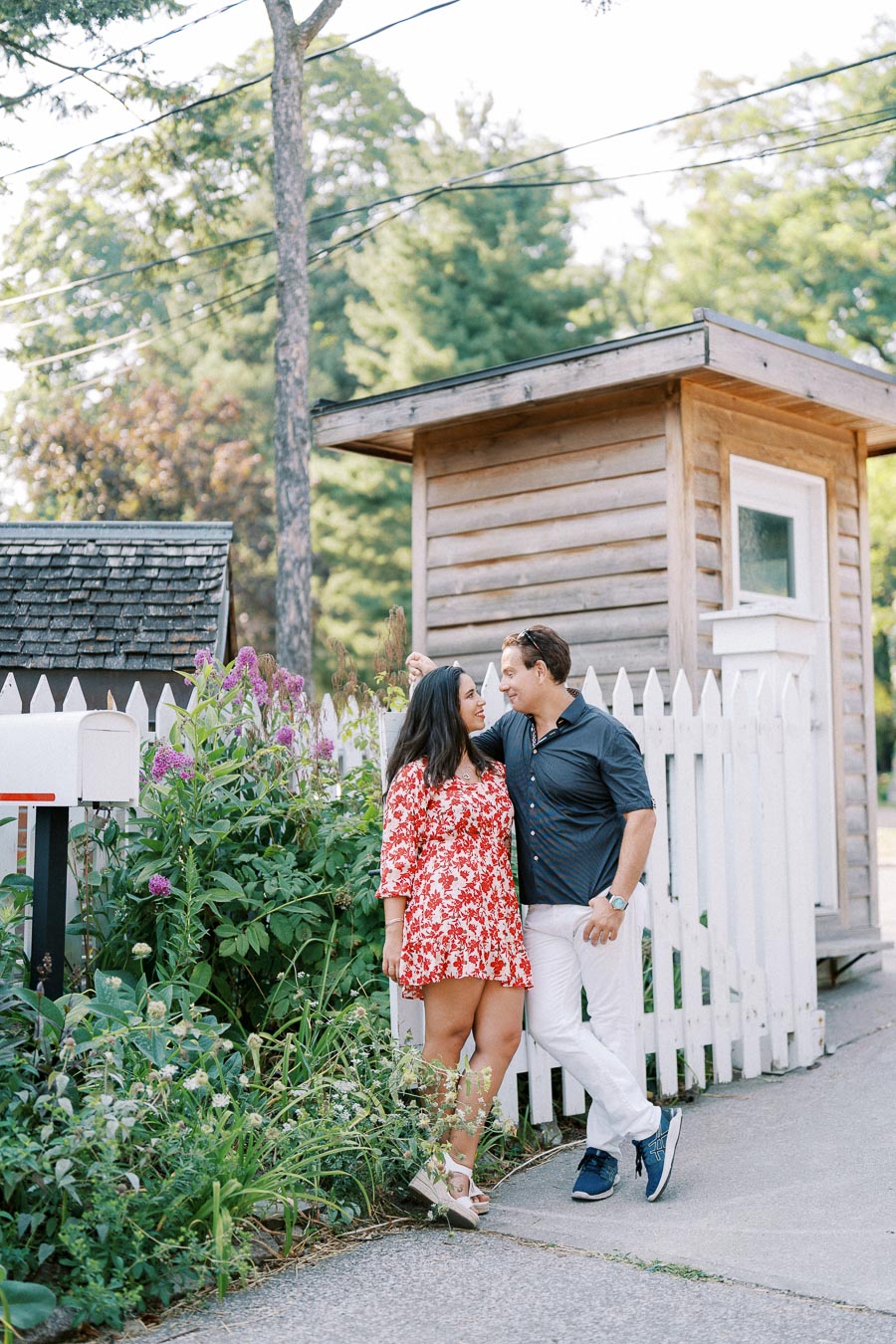 Couple standing by a rustic wooden shed and white picket fence, with flowering plants in the foreground, set in a lush, green outdoor environment. The woman is wearing a red floral dress, and the man is in a dark shirt and white pants.