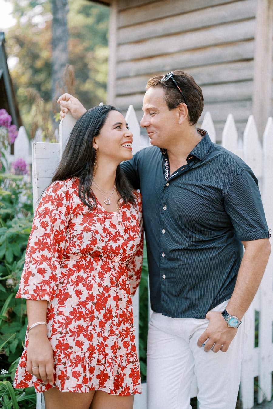 A happy couple smiling at each other in a garden setting, with a white picket fence and blooming flowers in the background. The woman is wearing a red floral dress, and the man is in a dark shirt with white trousers.