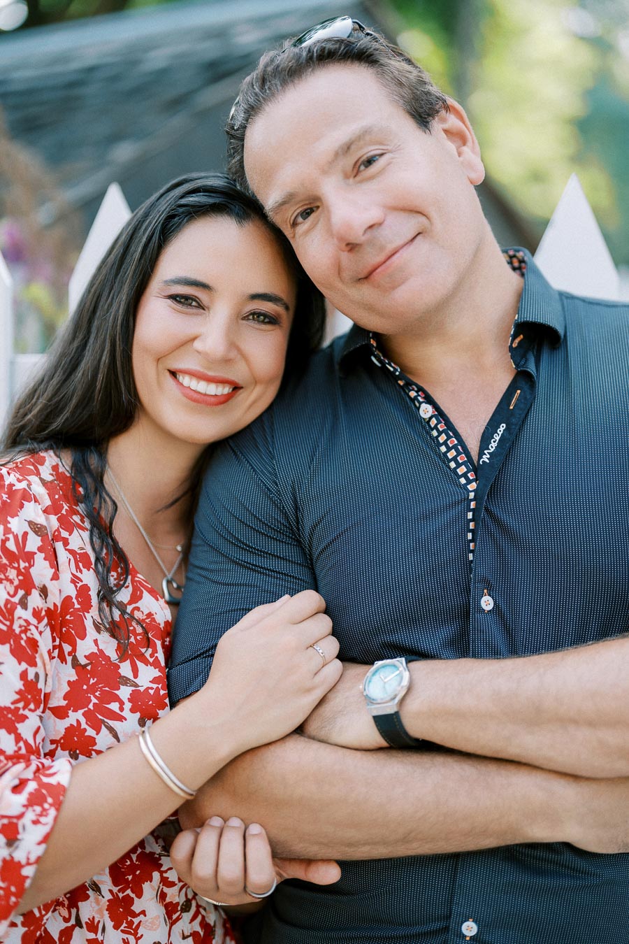 A happy couple embracing in front of a white fence, with the woman in a red and white floral dress and the man in a navy blue shirt, both smiling warmly at the camera.