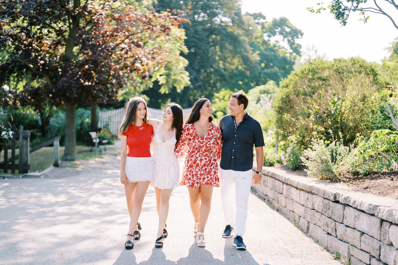 A group of four people walking together on a sunny day in a park, with lush green trees and a stone wall in the background. They are all smiling and wearing casual summer clothing.