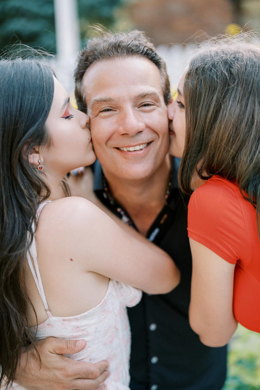 A happy man receiving kisses on each cheek from two smiling women, outdoors in a joyful, family moment.