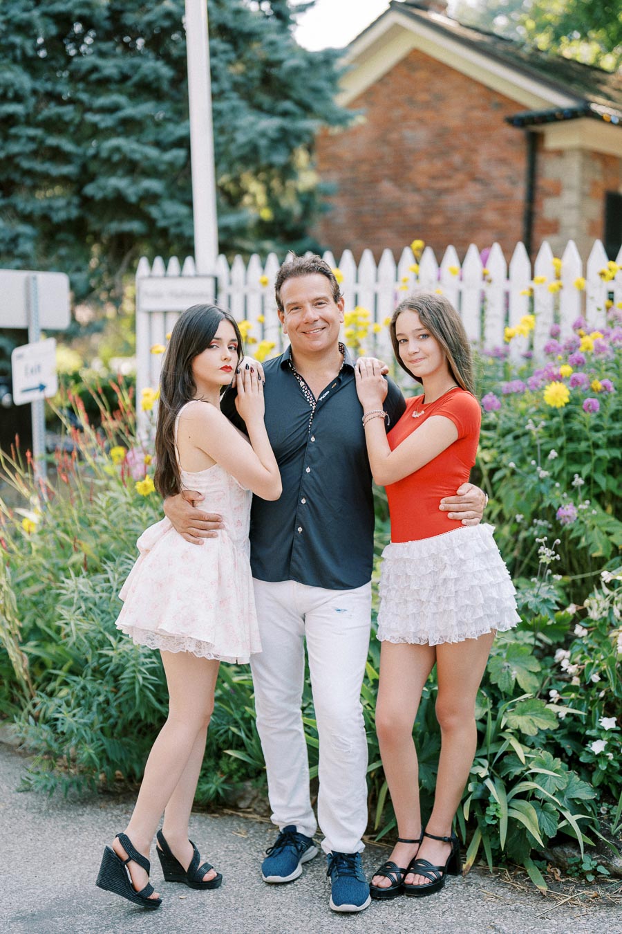 A man standing between two young women in a garden setting with colorful flowers and a white picket fence in the background.