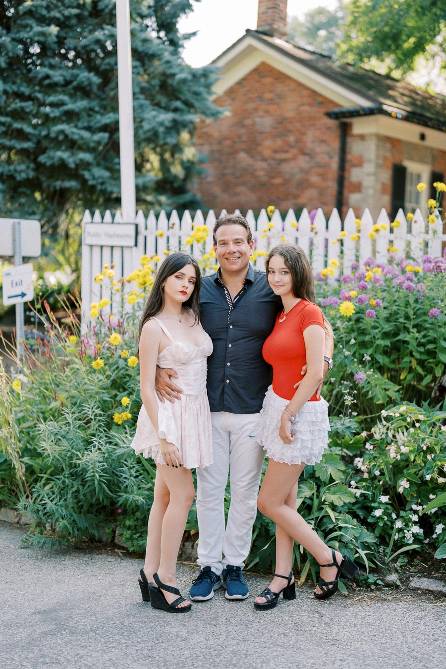 A group of three people smiling and posing in front of a colorful garden with a white picket fence on a sunny day.